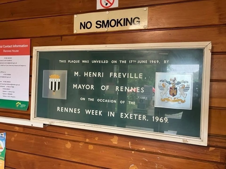 A wall plaque saying it was unveiled in 1969 when the Mayor of Rennes visited the tower block. It's mounted on a wall clad in wood. Above it someone has used drawing pins to stick up a no smoking sign.