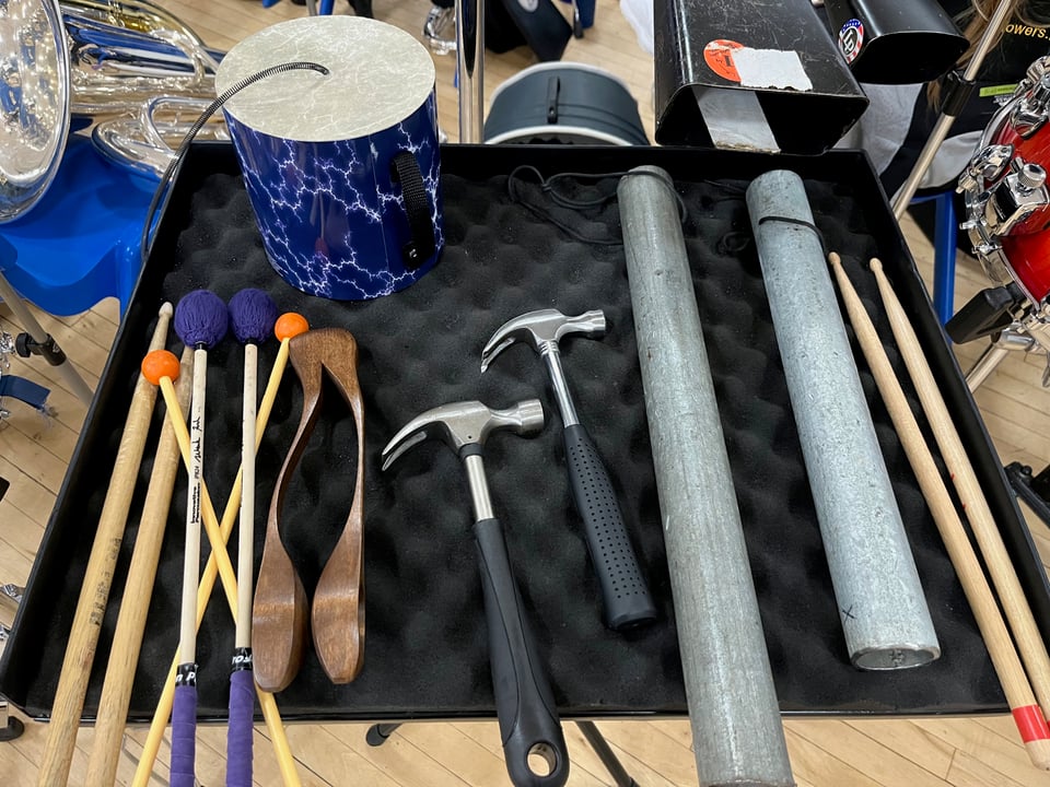 A collection of percussion tools laid out on a table. From left to right: several pairs of drumsticks, some sort of wooden castanets-like thing, a pair of hammers, two scaffolding poles, and two more drumsticks. There‘s also a large blue cylinder with a black spring coming out the top at the top of the table. Behind the table you can see other brass instruments.