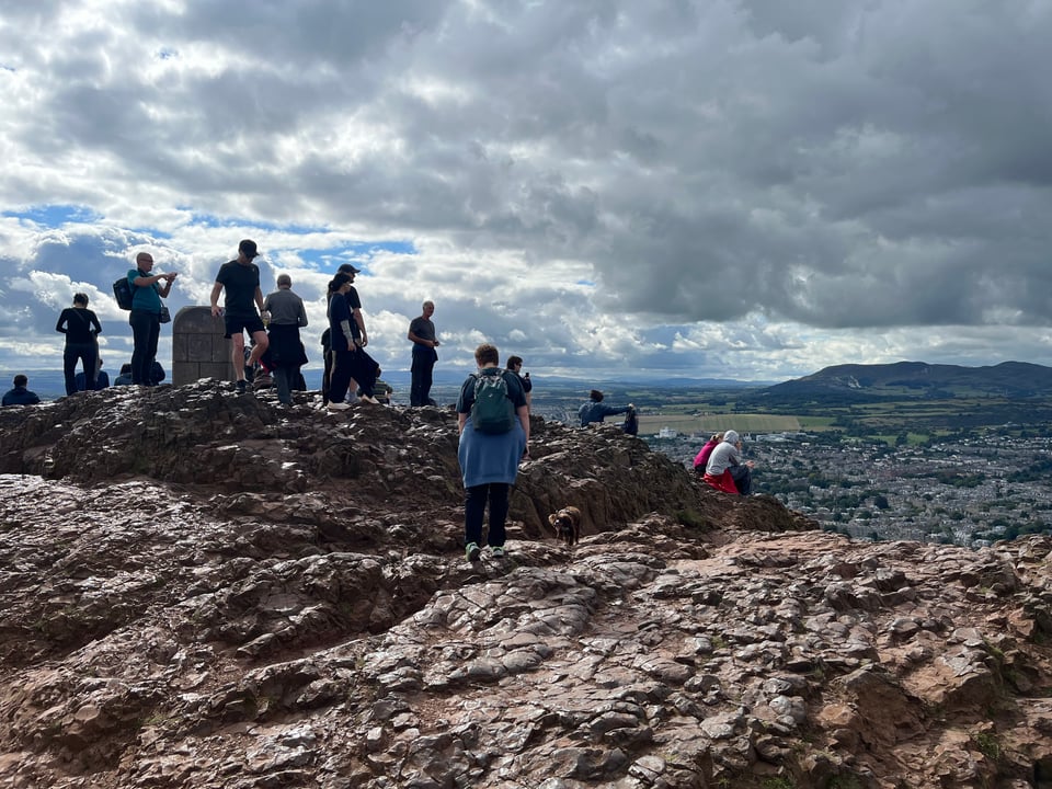 A photograph of several people standing on a rocky summit, overlooking a sprawling city in the distance, under a heavy gray sky.