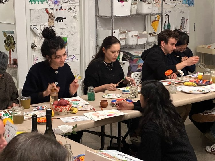 An instructor crouches at a table where students are painting watercolor still lifes