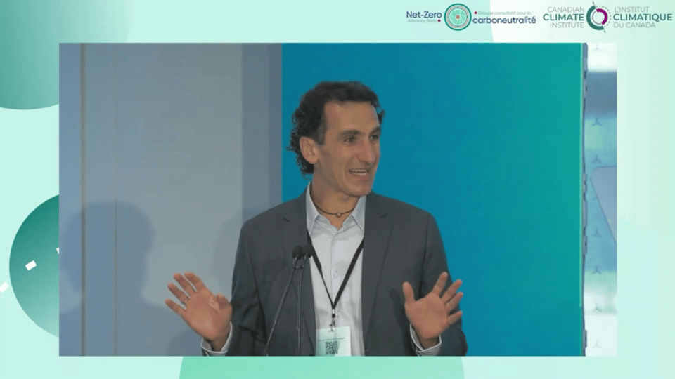 a clean shaven middle aged white man dressed in a pale blue shirt and grey suit, speaking to an audience at a conference. He is smiling and looking to his left.