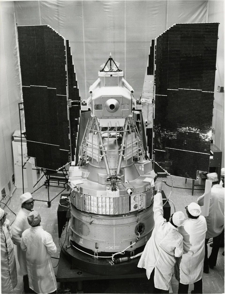 A large, silver metal cone with mesh and wires and four large black panels coming off it like elephant ears. A group of people in white lab coats inspect it. This is Landsat-1