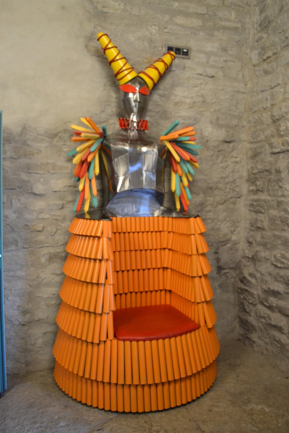 A chair where the person sits in the skirts of a figure  wearing armor, colorful "feathers" on the arms, and a jester hat.