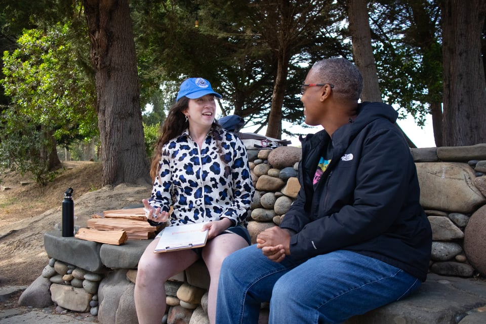 Hans and Saifa sit outside at a fire pit made of large stones, next to a pile of firewood. Hans is holding a clipboard, talking, and gesturing, as Saifa clasps hands and listens.