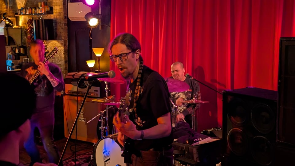 A three piece band (guitars, drums, and bass) plays in front of a red curtain. In the center of the frame, the bassist and singer, Jay, is dressed in black wearing glasses and passionately playing and singing. He has a long braid that hangs over one shoulder. The guitarist and drummer are also playing passionately.