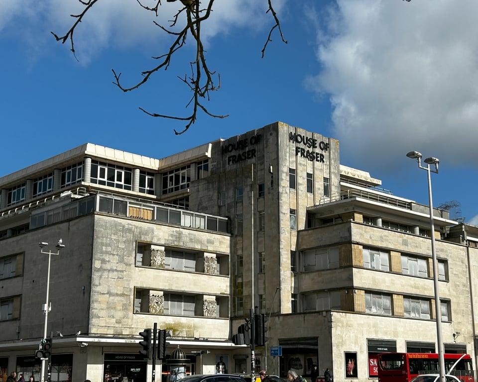 A 1950s department store on a sunny winter's day in 2025. The building is made of two horizontal blocks joined by a taller tower. There are ribbons windows and some kind of rooftop terrace. all the stone is discloured by pollution and there is moss or lichen growing on some of it.