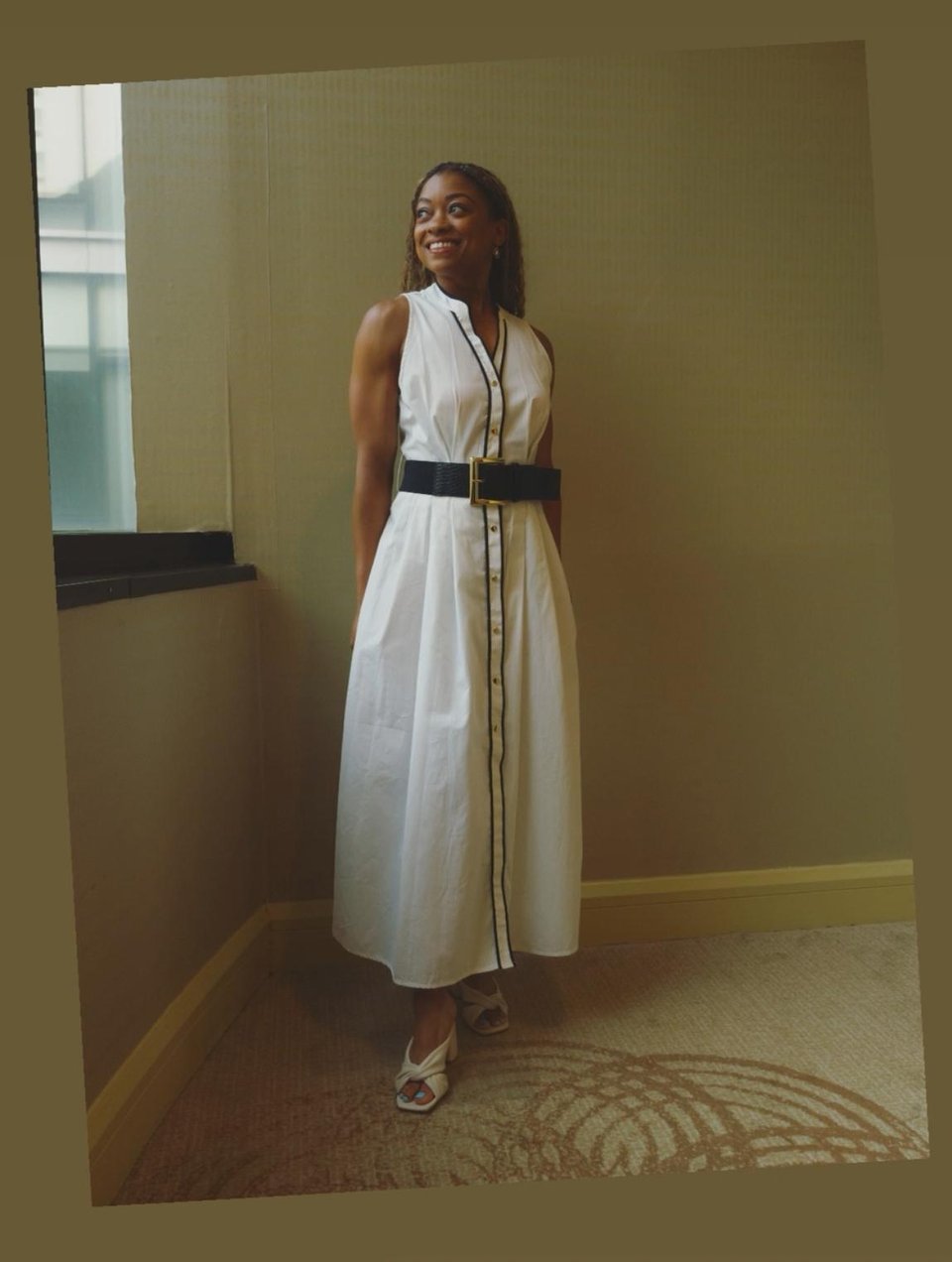 I’m standing by a window in a softly lit room, wearing the same white sleeveless dress with black piping and a bold black belt. I’m looking off to the side with a bright smile, my hands tucked into my pockets, and the hem of my dress just skimming my ankles. I'm wearing white crisscross heels.