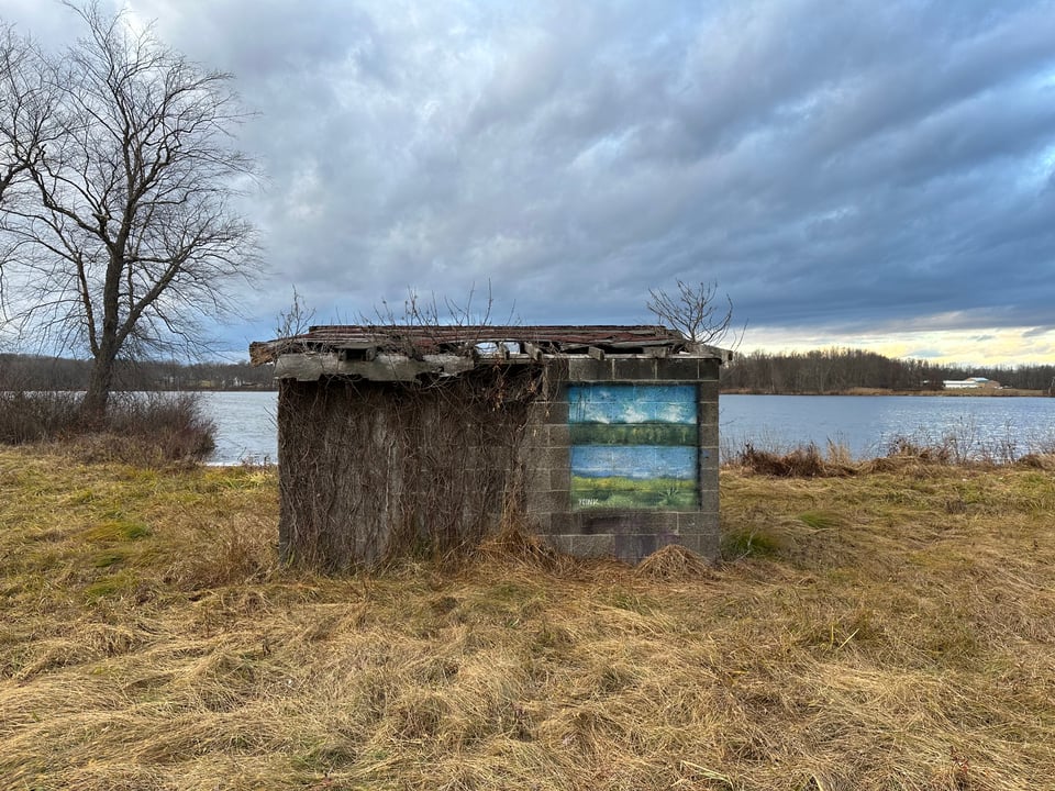 photo of concrete bunker with rotting wooden roof by lake; the bunker has a lakescape painted on it like a window