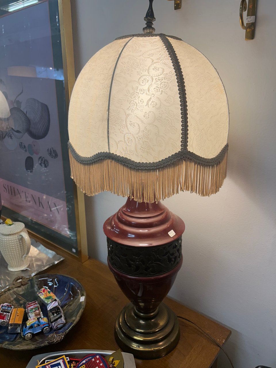 a cream-colored fringed lamp with an oxblood red base sits on a wooden table.