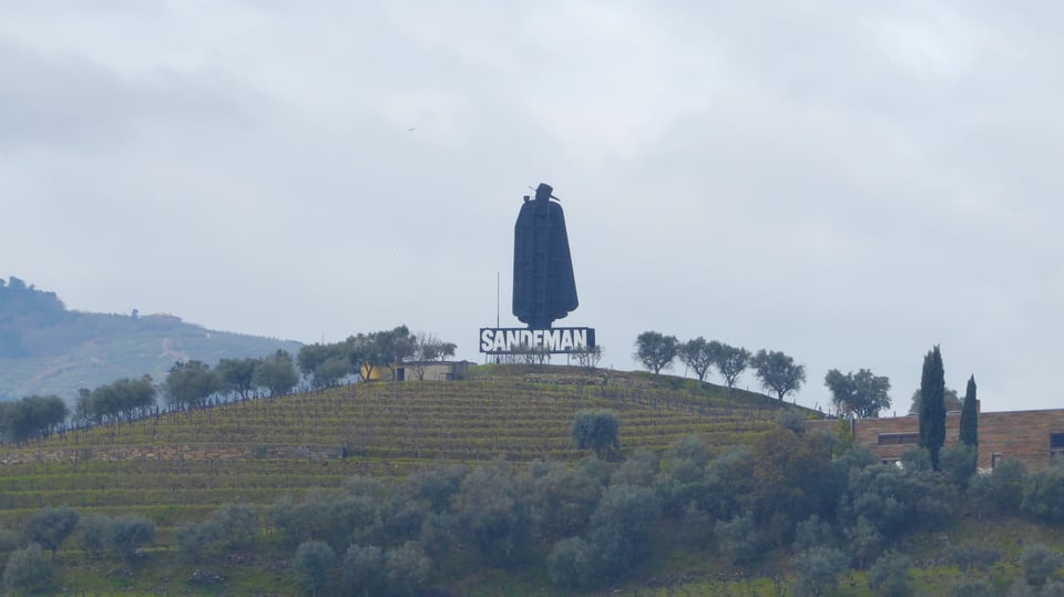 Photograph of a terraced vineyard on a hillside. On the top of the hill is a large purpose-built structure in the shape of a man in a cape and hat. Underneath the man is a large sign that says, "SANDEMAN"