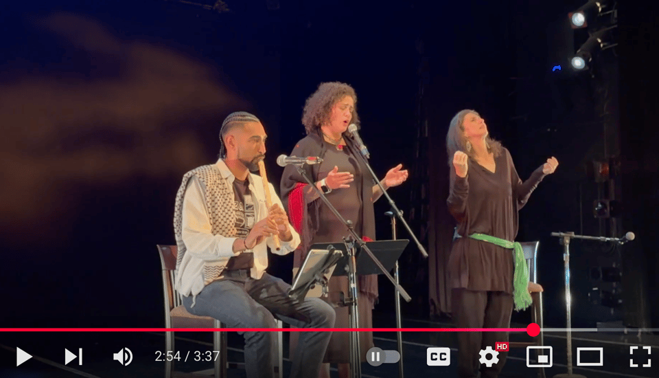 Image description: A still from a concert with three people on a dark theater stage, one with a flute, one singing, one dancing