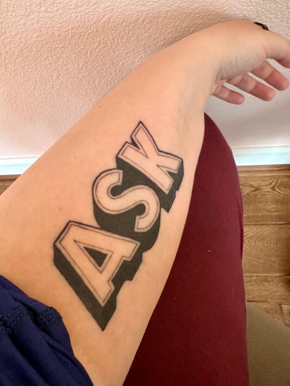 A white forearm resting on burgundy pants. The word "ASK" is tattooed in sign painter Gothic lettering with a dramatic drop shadow across the forearm.