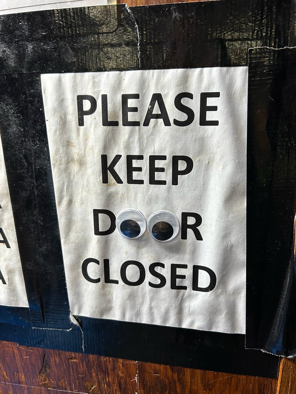 A white sign with black says "PLEASE KEEP DOOR CLOSED" and is taped to wood paneling (ON A WALK IN?) with big googly eyes over the OOs in "Door."