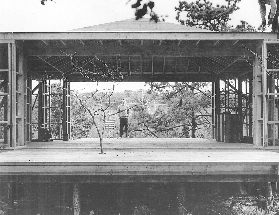 A black and white photograph of the wood frame of a house under construction. A man stands on the far side of the house.
