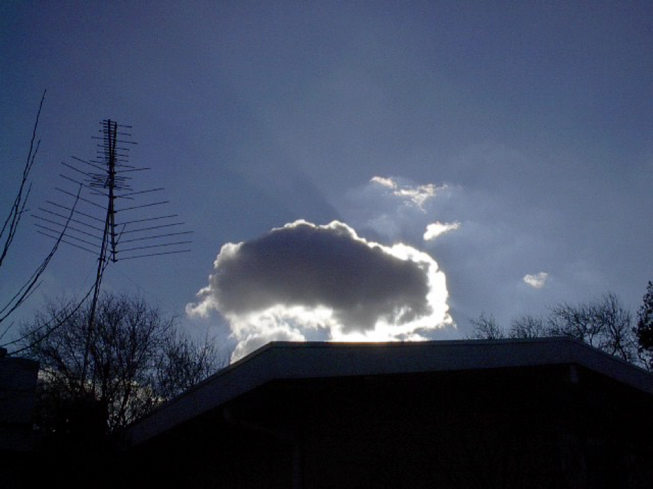 a low-resolution image of a cloud, backlit by the sun in an otherwise blue sky, over the silhouette of suburban houses, trees, and an antenna