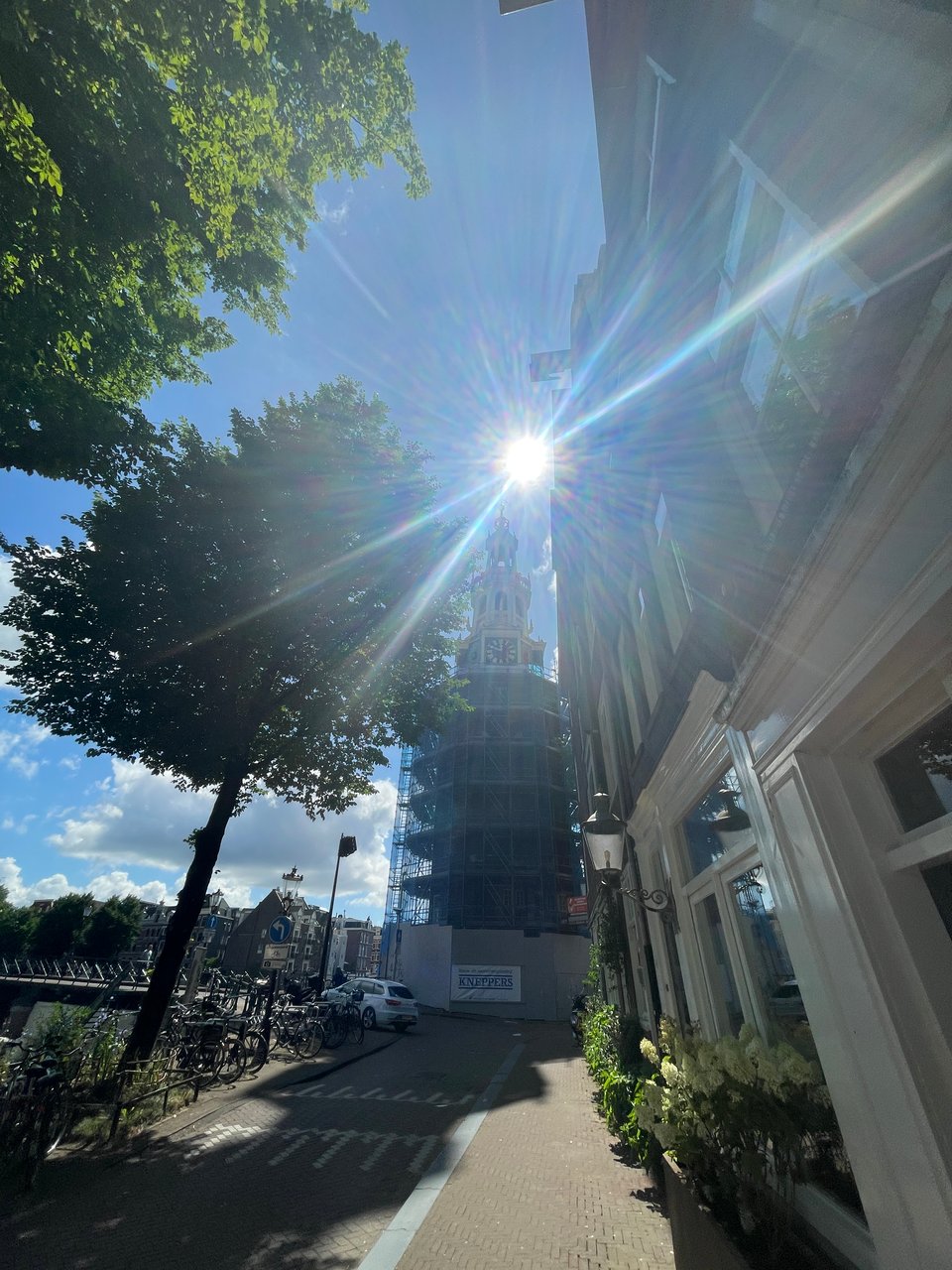 A sunny street in Amsterdam ends in a bell tower under scaffolding for repairs.