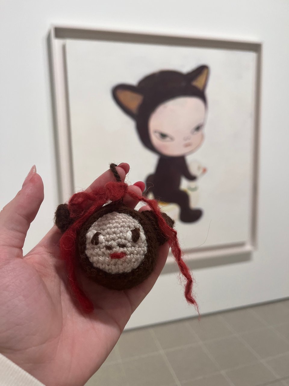A photograph of a painting called Harmless Kitty by Yoshitomo Nara. It shows a young child in a brown cat onesie, sitting on a duck as though it was a horse. The child faces away from the viewer, but looks back over their shoulder. The background of the painting is white. In the foreground of the photo, Kel's hand holds up a small crochet charm of the character's face. It also has a red ribbon on top.