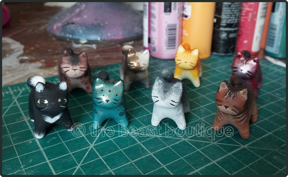 A collection of eight tiny cat sculptures in assorted colors. (Tuxedo, Brown, Pthalo Green, Metallic Calico, Grey, ORange Tabby, Pink Tiger, and Brown Tabby)