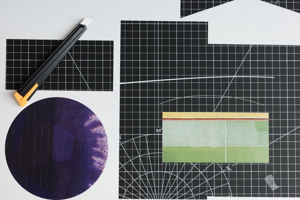 Studio work surface showing black cutting mats with grid lines, a rotary cutter with orange handle, a circular deep purple textile piece with vertical stitching, and a rectangular color-blocked composition in sage green and yellow-green tones with thin red accent line
