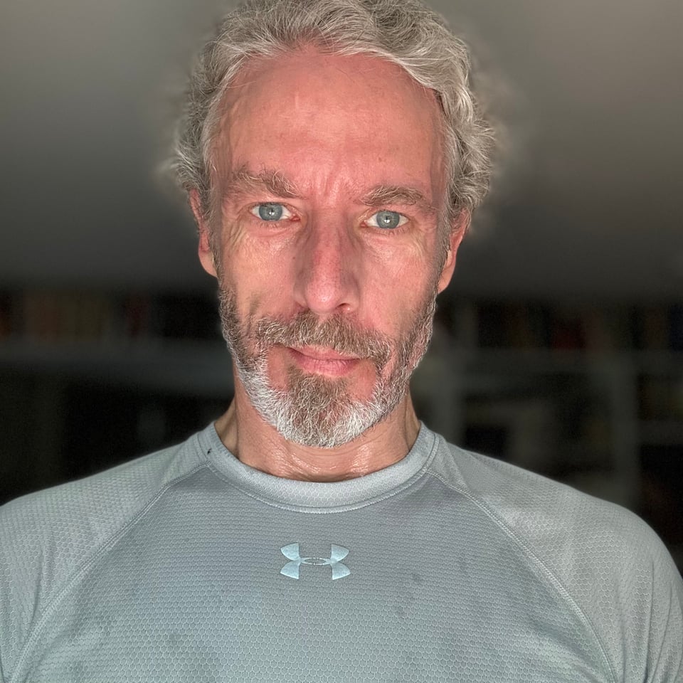 Photo of a white man with a flushed face semi-smiling at the camera, wearing a running shirt that is doused in sweat.