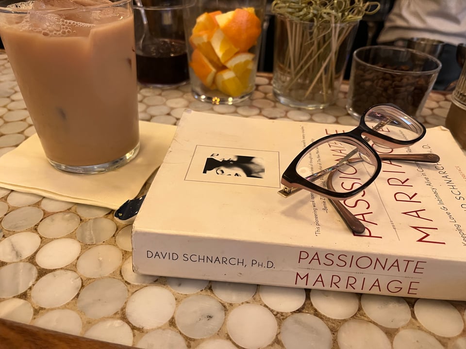 close-up of a bar made of small round stones, with a glass filled with milky tea and the book Passionate Marriage with glasses sitting on top. in the background are glasses filled with cut oranges and other bar suppliees.