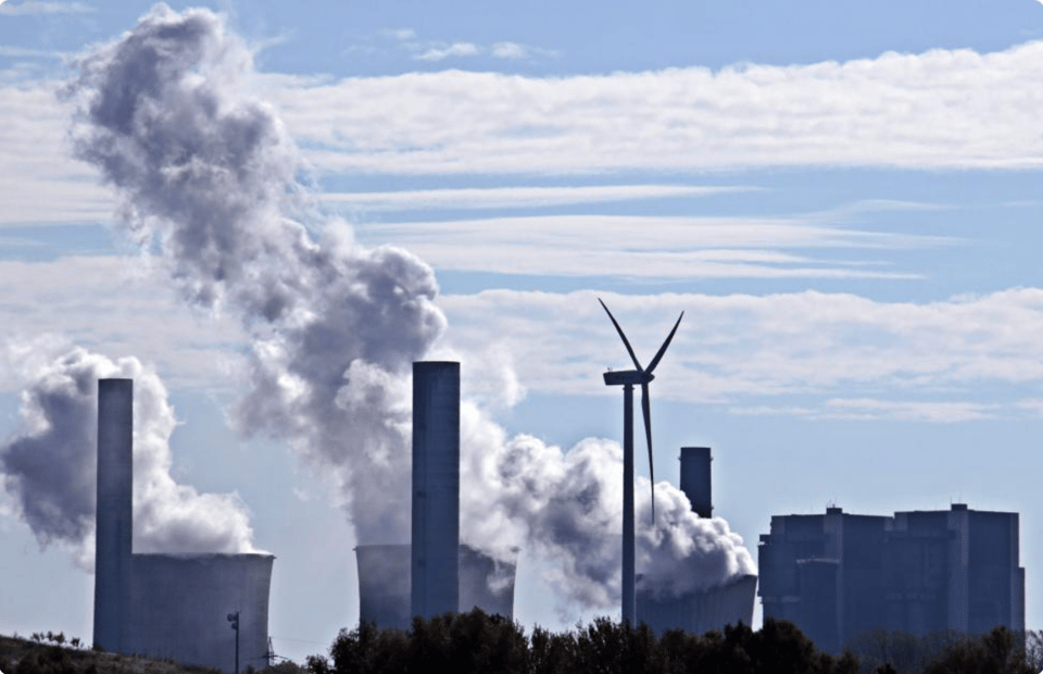 Photo of smoke stacks, maybe from a power plant, with white clouds billowing out of them plus one lone windmill.