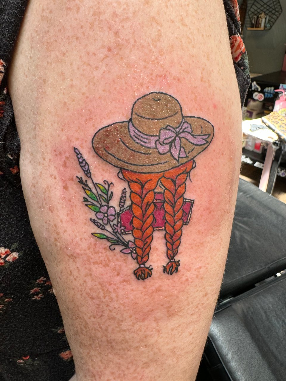 A tattoo on fair, freckled skin, showing a straw hat with a purple ribbon tied in a bow at the front, 2 bright red-orange braids extending from beneath it, and lavender, violets, and green leaves scattered around the image