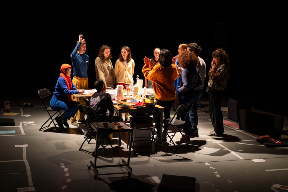 A group of people are clustered around a table full of items, lit by stage lighting. Some are gesturing as the group discuss.
