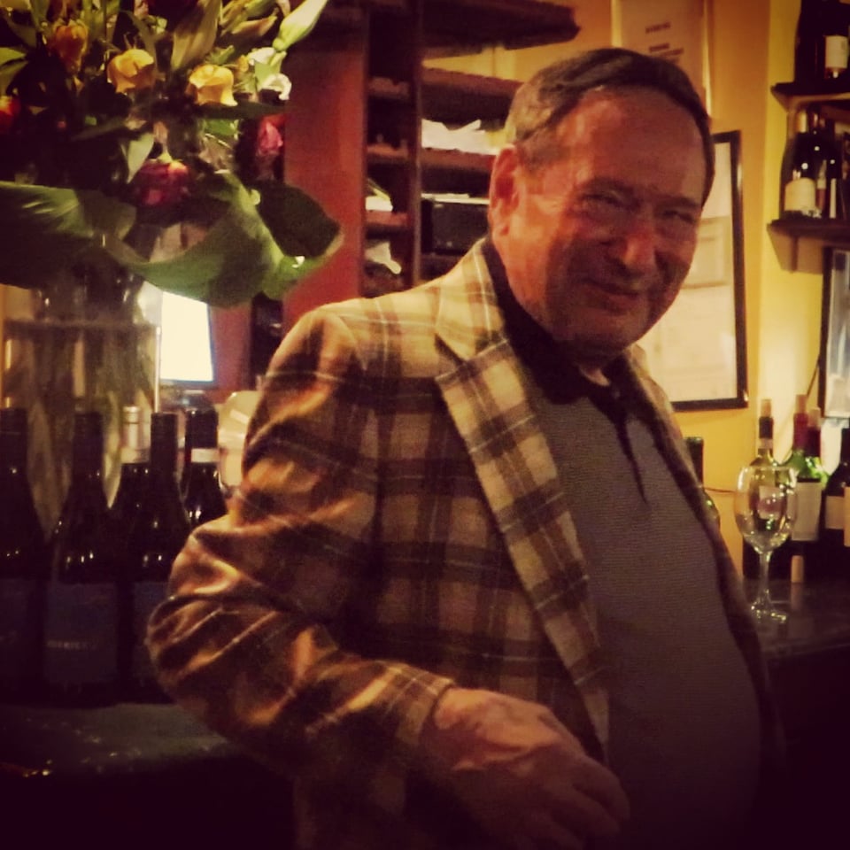 Man in plaid jacket smiling next to bottles of wine in restaurant