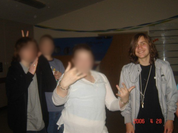 Four teenagers stand in a poorly lit multi-purpose room. One is holding his hands up doing a karate pose, while a friend behind him gives him bunny ears. One has her hands raised, middle finger down. On the right, Kurt is visible, with shoulder-length hair ending in ringlets. He wears a black t-shirt and a grey jacket. His keys are worn around his neck. A date reads 2006 4 28.