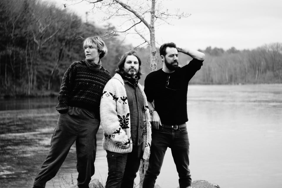 A black-and-white image of three musicians standing on the shore of a lake, each looking in a different direction.