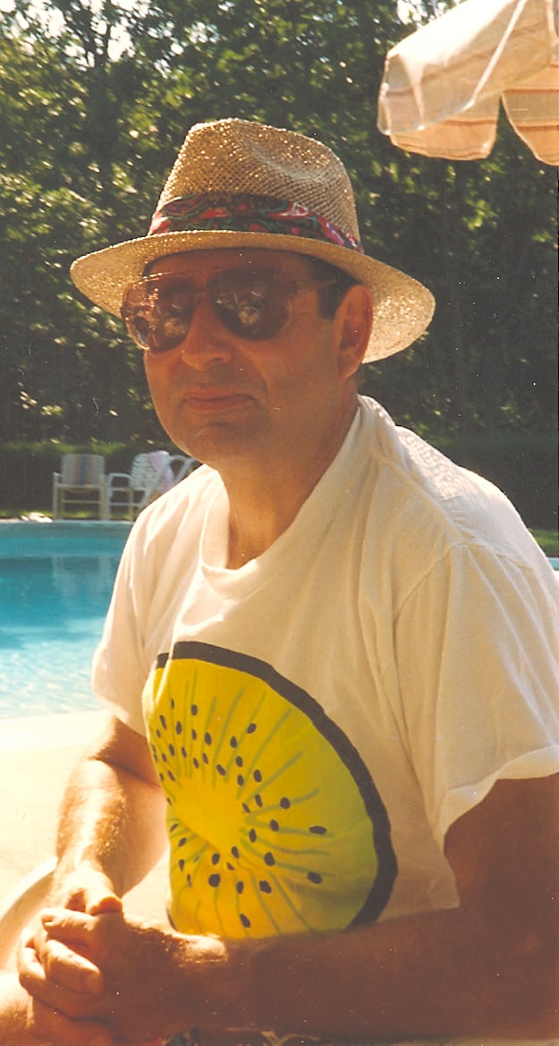 Man in straw hat and sunglasses wearing white shirt printed with large kiwi, sitting next to pool on sunny day