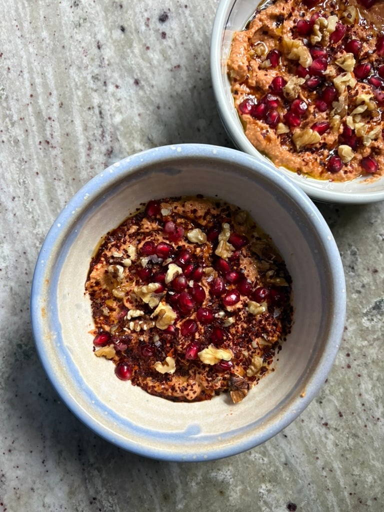 A blue and tan bowl of red pepper dip muhammara topped with chopped walnuts, pomegranate seeds, Aleppo pepper flakes, olive oil, and flaky salt.