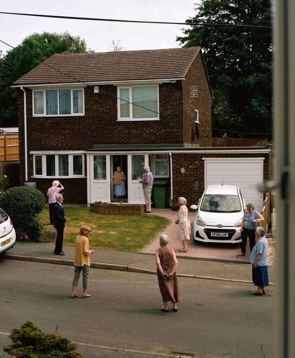 Photograph of elderly neighbours standing in small groups on a suburban street and driveway outside a brick house, with a couple at the open front door
