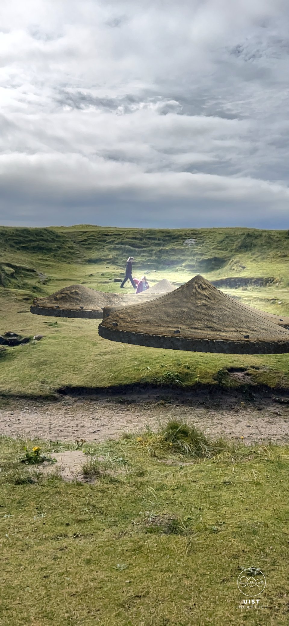 A view of the grassy, hilly landscape and roundhouse of Cladh Hallan but with an approximation of the original conical turf roofs superimposed by augmented reality. It gives an idea of what the roundhouses originally looked like.