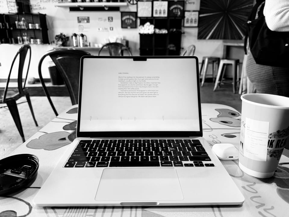 b/w photo of an open laptop on a table in a coffee shop with a coffee cup and an AirPods case next to it