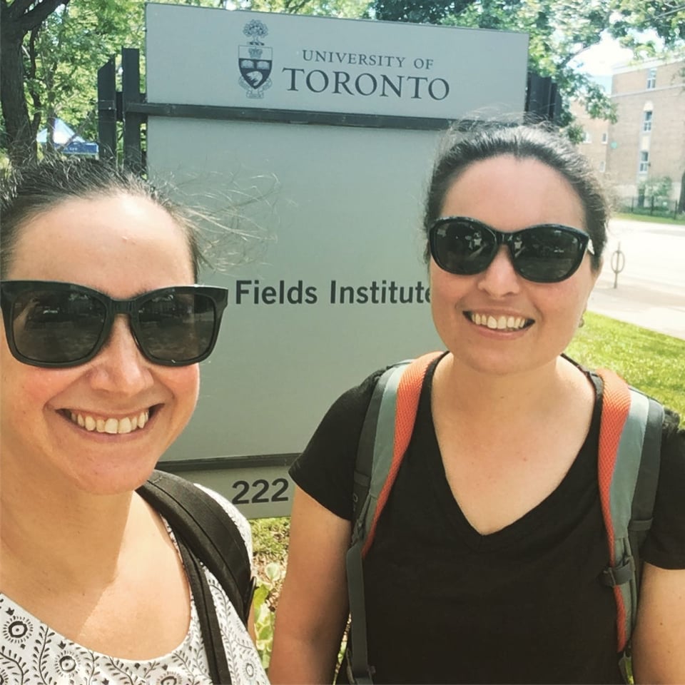 Two people with matching sunglasses, backpacks, pulled-back hair, and big smiles, in front of a sign that says University of Toronto, on a sunny day.
