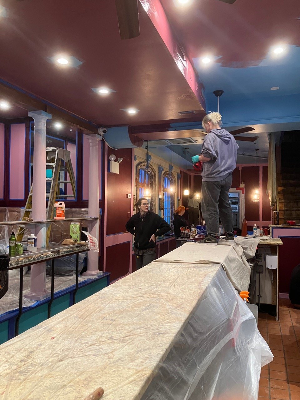 Co-owner Clover discussing painting the ceiling above the bartop with a friends who is standing on the covered bartop, ready to paint. The bar is partially the original bright blue, and partially the Val's red.