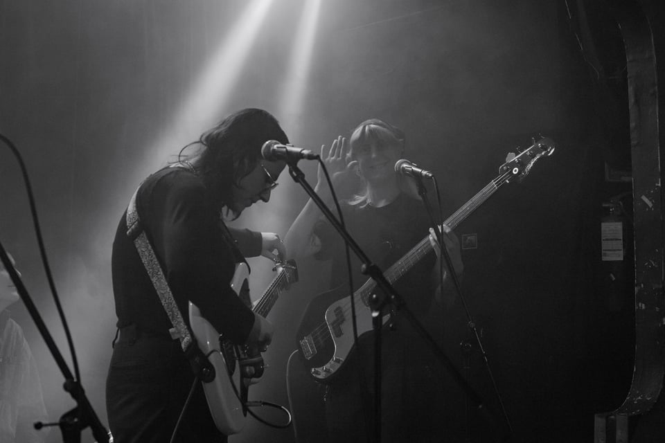 lotus, holding the bass, waves at the camera while claire faces away tuning