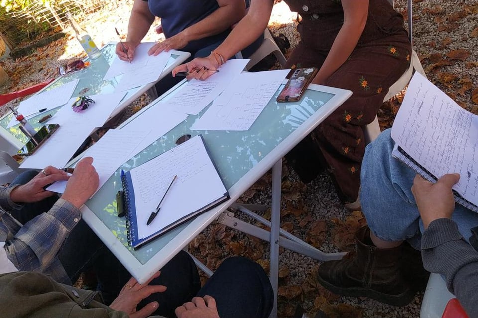 cinq personnes dans un petit jardin en train d'écrire autour de deux tables couvertes de papiers et stylos