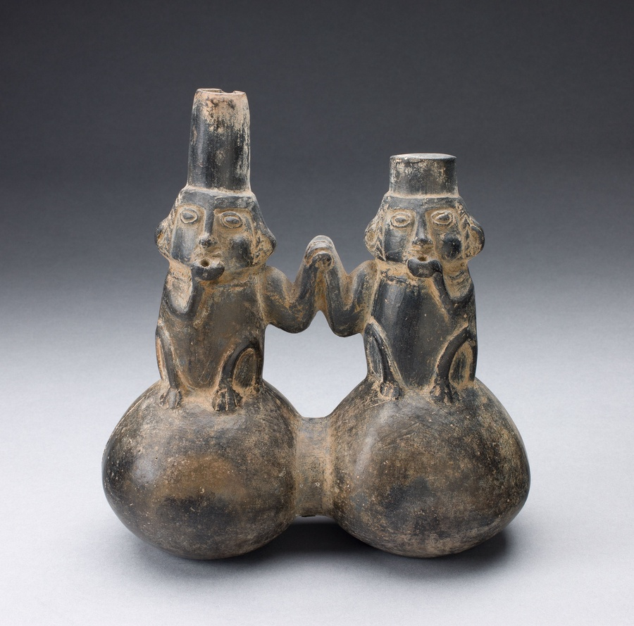 A dark colored ceramic vessel in the shape of two people holding hands. Their bulbous lower bodies are joined at the base of the vessel and they wear cylindrical hats that appear to be spouts.