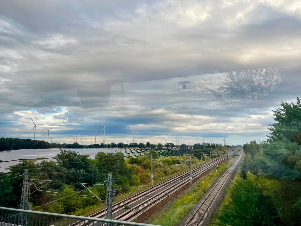 Train tracks lead to the horizon. On the left a field full of solar panels stretches off into the distance and windmills dot the horizon.