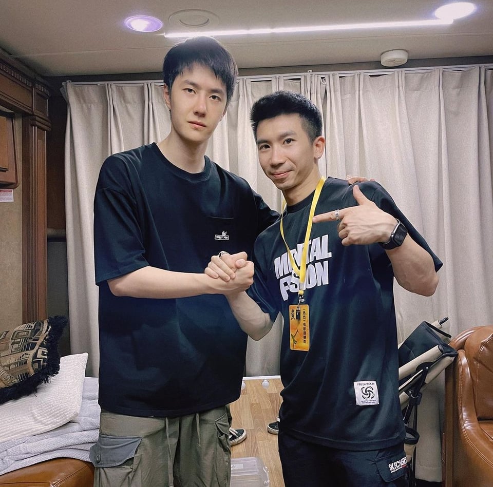 picture of Wang Yibo standing in what looks like a hotel room next to a shorter guy. They are clasping hands in a "bro" sort of way and the shorter guy is pointing at Yibo, who has his hand on the other guy's shoulder.