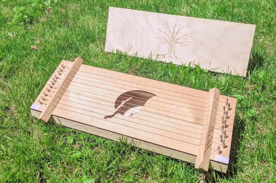 An aeolian harp lying in the grass. It is made of light pale wood, and looks like a short rectangular box perhaps two feet long and one foot wide. It has a dozen metal strings, and a soundhole in the shape of a leaf