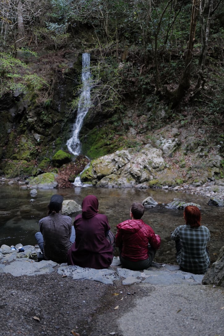 Four people seated side by side on flat rocks at the edge of a still pool, quietly watching a slender waterfall thread down a moss-covered cliff face through dense forest