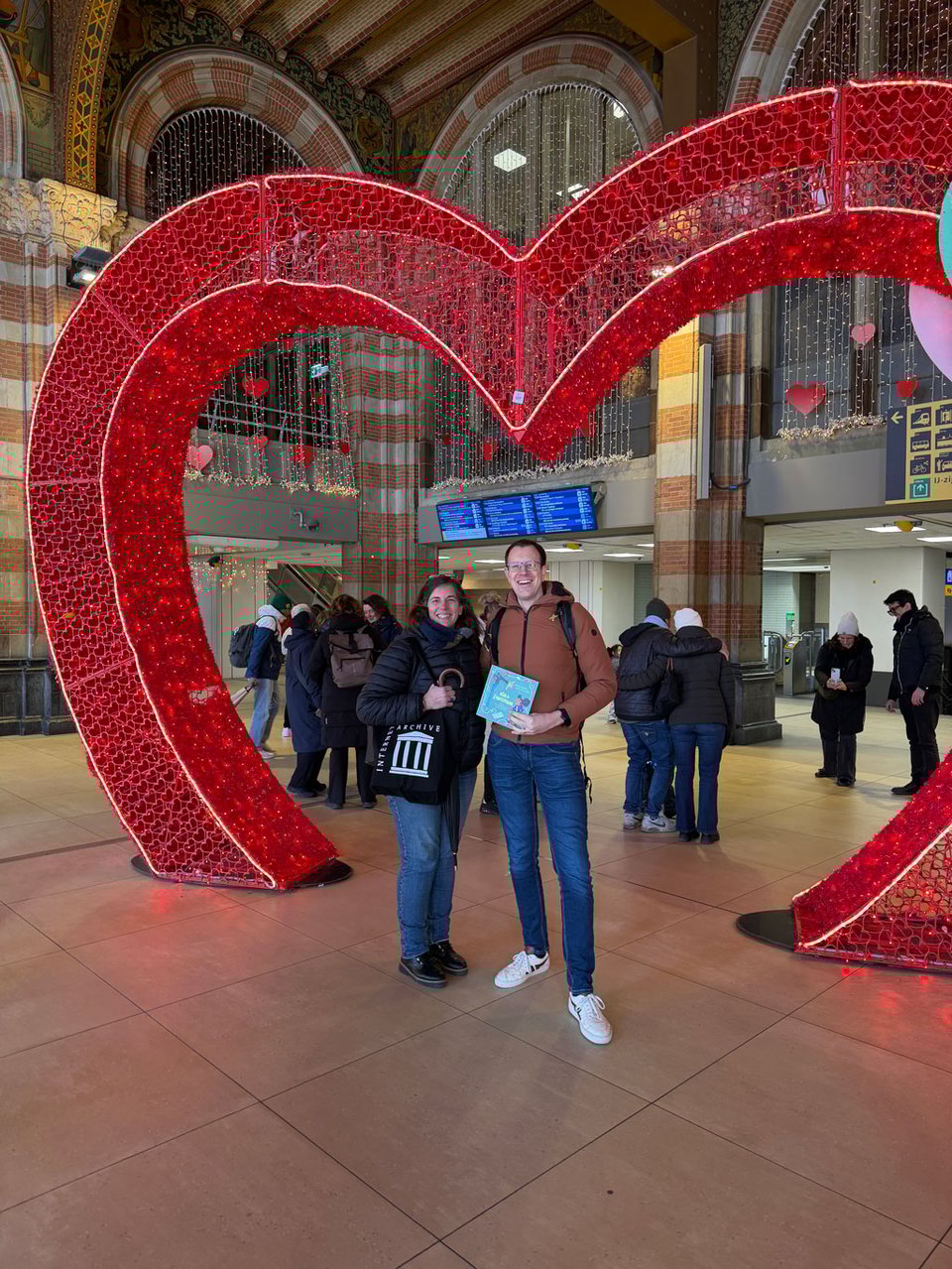 A woman and a man stand under an electric lit up heart. She holds and Internet Archive tote bag and he holds the book “Ada & the Zangemann”.