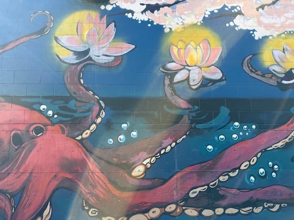 Mural painting on a brick wall of a red octopus in dark blue water holding three glowing pink lotuses in its arms beneath of a blossoming cherry tree
