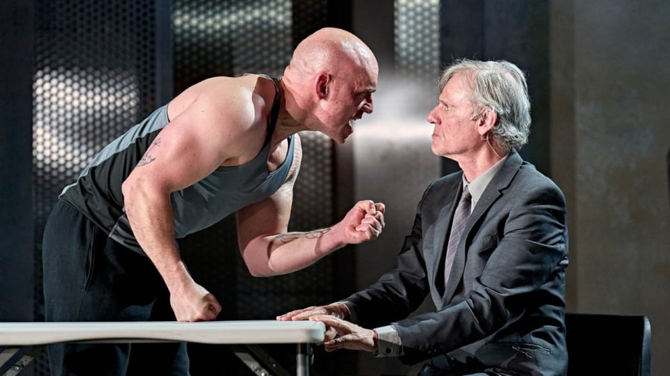 Samuel Edward-Cook as Raoul Moat, and Trevor Fox, in Manhunt. Fox is wearing a grey suit with a grey shirt and grey tie; he has grey hair. He's seated at a trestle table. Edward-Cook is in front of him, getting in his face, red-faced and fist curled-up. He is wearing a grey gym singlet and black trackpants.