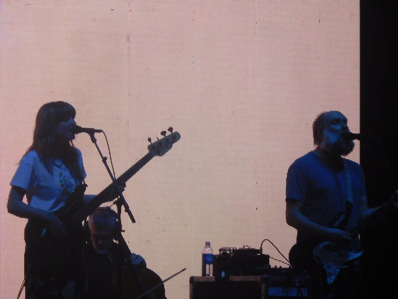 Two people (Melanie Radford and Doug Martsch) singing and playing electric bass and electric guitar, respectively, on a festival stage.