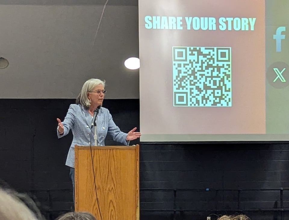 Katherine Clark stands behind a podium with her arms outstretched as she makes a point about what we need to do to take action.